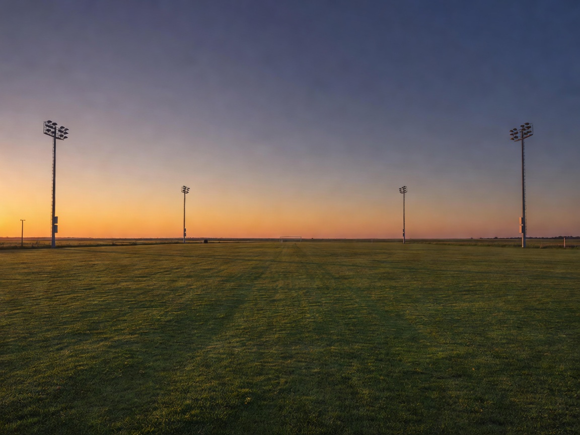 Evening light falling across an open sports ground with an empty outfield and floodlight towers in the distance
